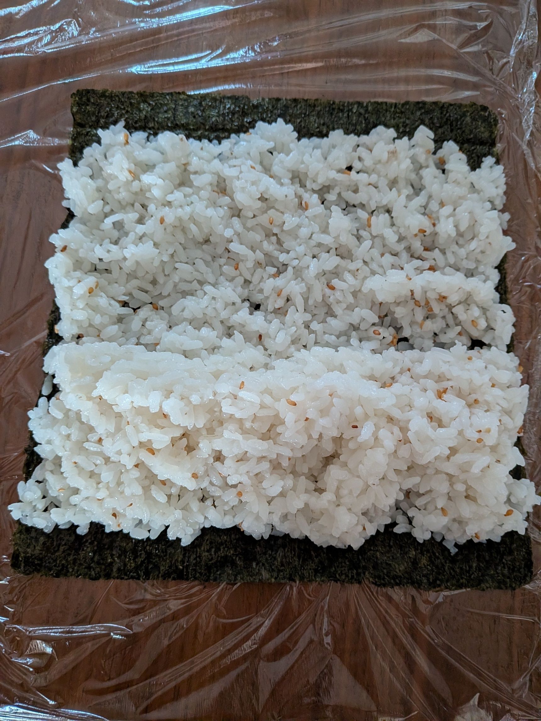 2、海苔をラップの上に広げ、酢飯を広げる。少し手前をもりあげる。だいたい1本につき、300gの酢飯をのせる。